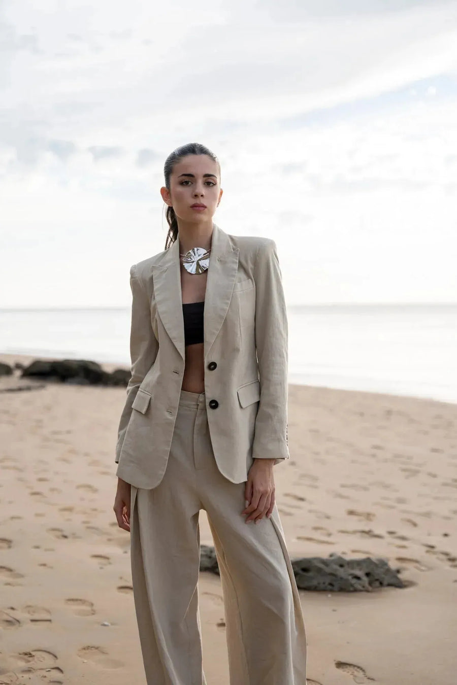 Femme sur une plage portant un pantalon large et fluide femme beige avec un bijou élégant, devant un paysage maritime.