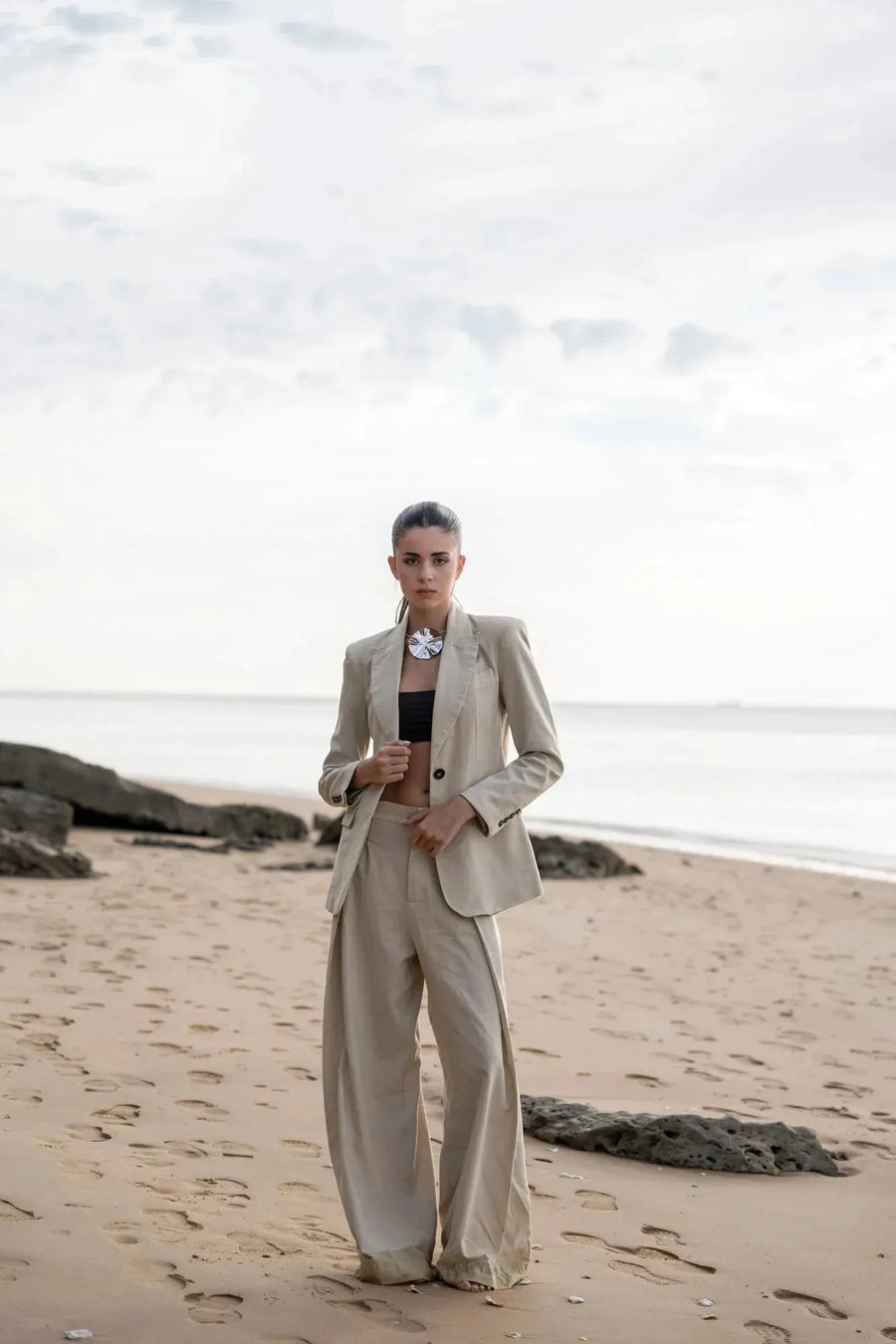 Portrait d'une femme en pantalon large et fluide femme beige sur la plage, tenant un blazer et affichant une allure moderne.