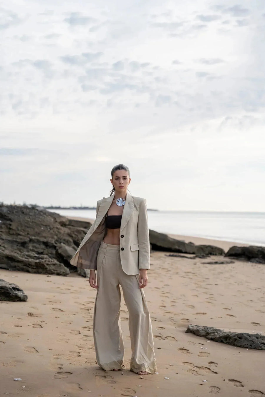 Femme élégante en pantalon large et fluide femme beige sur la plage, associée à une veste, style chic et décontracté.