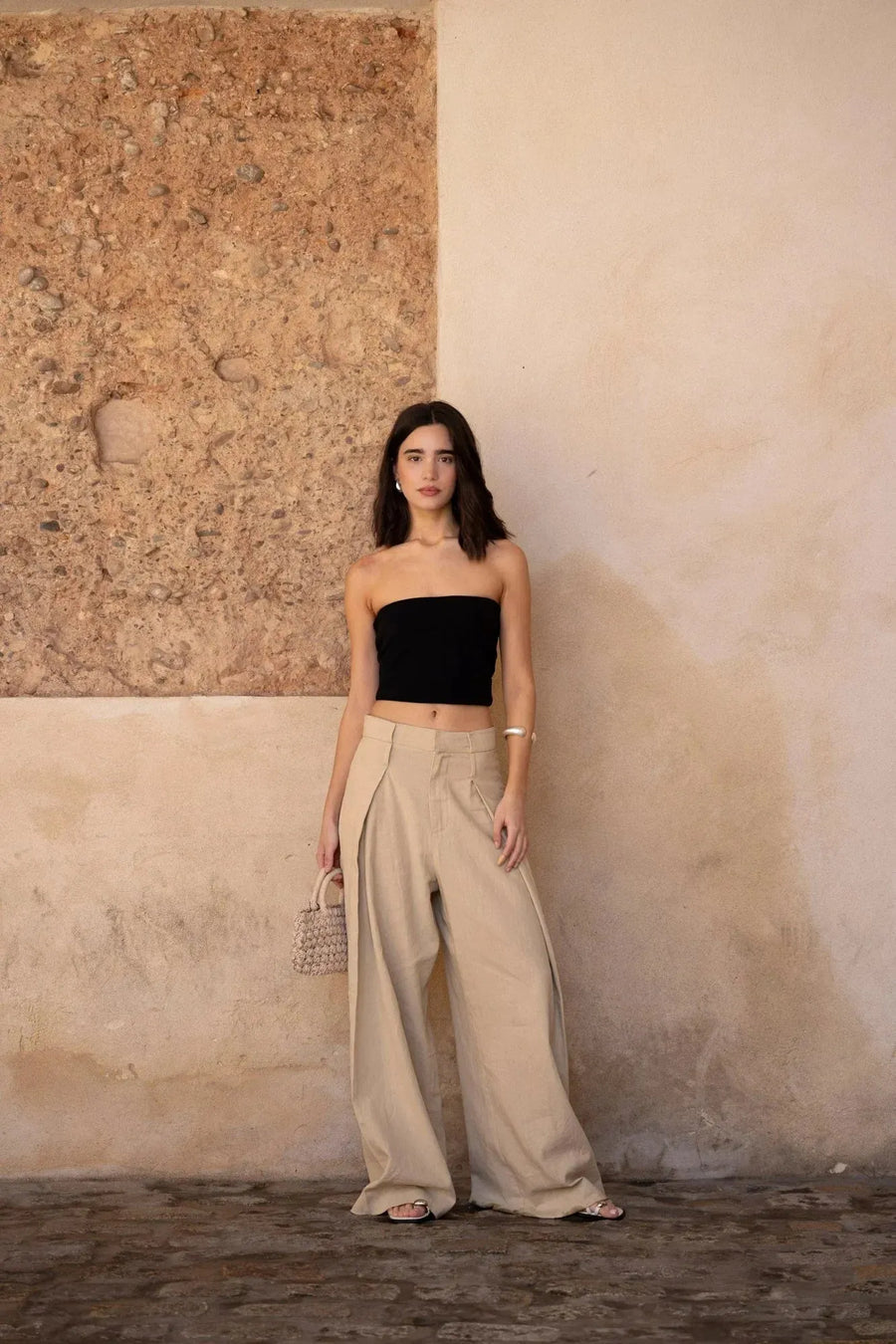 Femme posant avec un pantalon large et fluide femme beige, debout contre un mur en pierre, look décontracté avec un top noir.