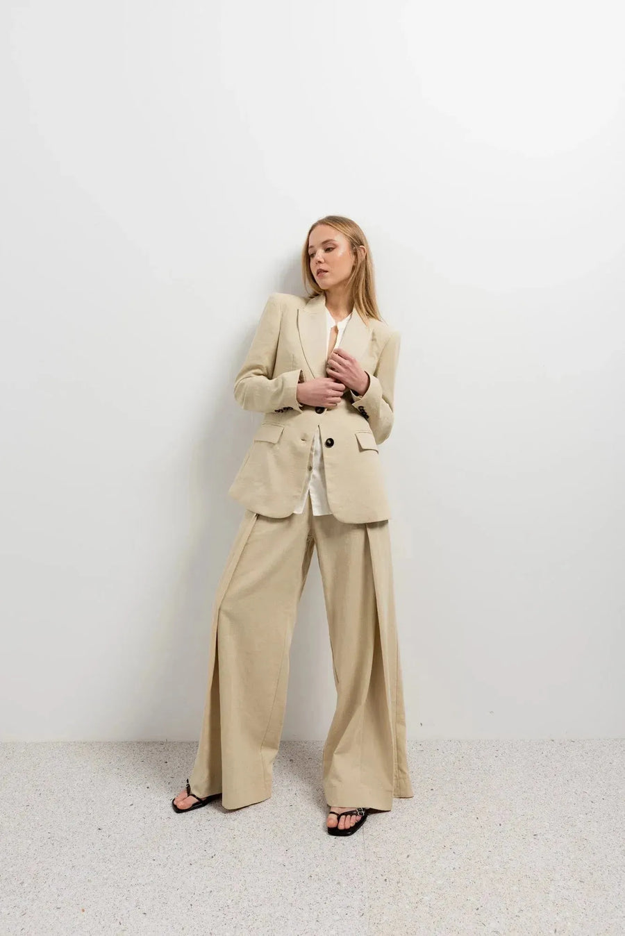 Femme posant avec assurance, vêtue d'un pantalon large et fluide femme beige et d'une veste associée dans un cadre minimaliste.