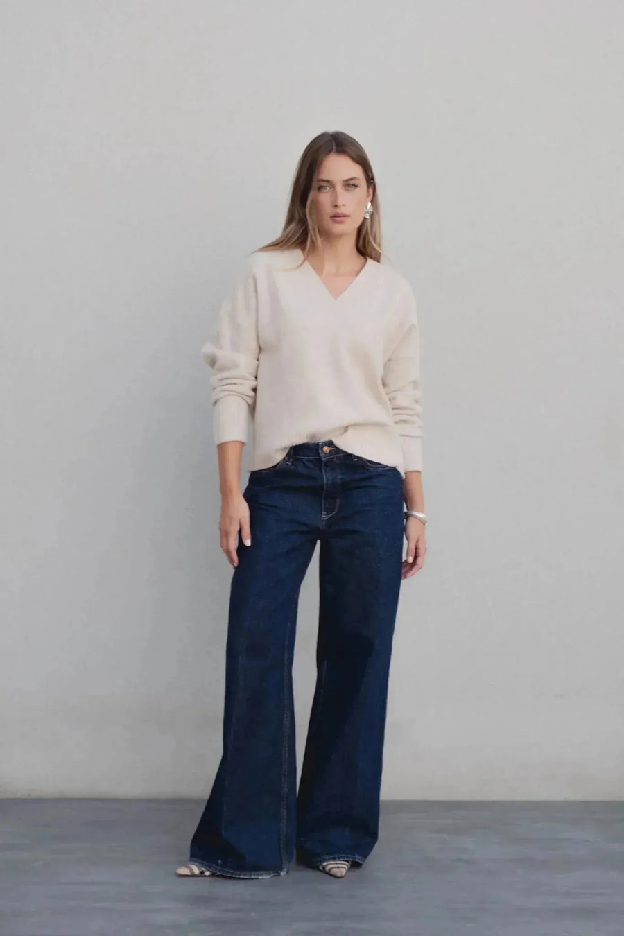 Mode femme avec un pull maille fine femme ample beige col V oversize, associé à un jean évasé, posant devant un fond beige.