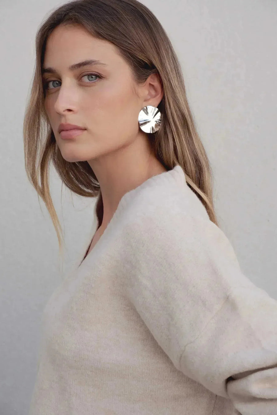 Femme montrant le pull maille fine femme ample beige col V, mettant l'accent sur son visage et ses boucles d'oreilles, avec un arrière-plan léger.