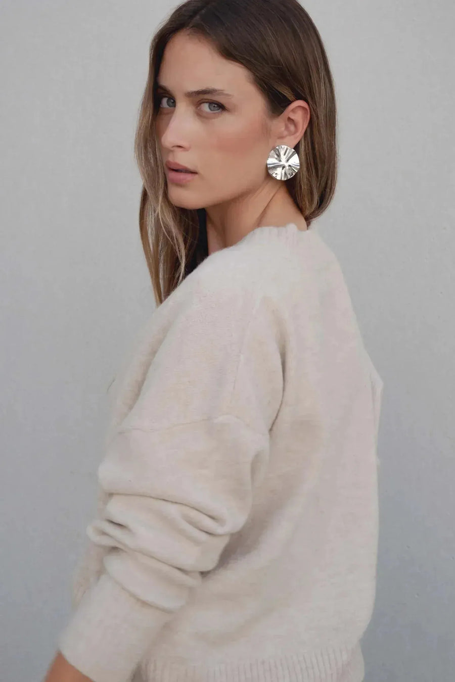 Gros plan sur une femme avec un pull maille fine femme ample beige col V, mettant en avant ses boucles d'oreilles brillantes, sur un fond neutre.