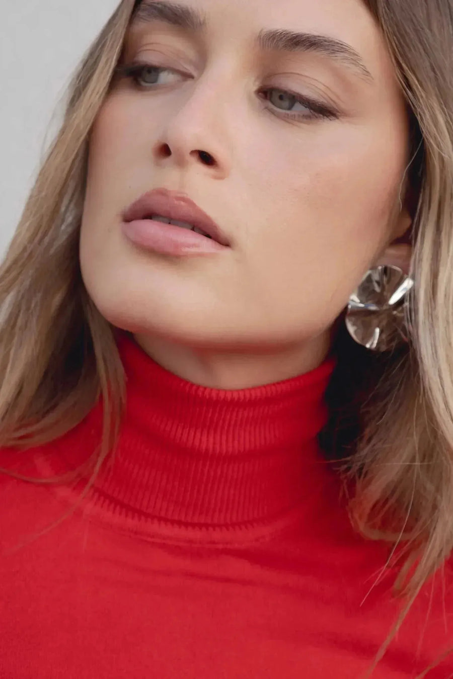 Gros plan sur un modèle avec pull col roulé femme rouge, affichant des accessoires élégants et une expression contemplative.