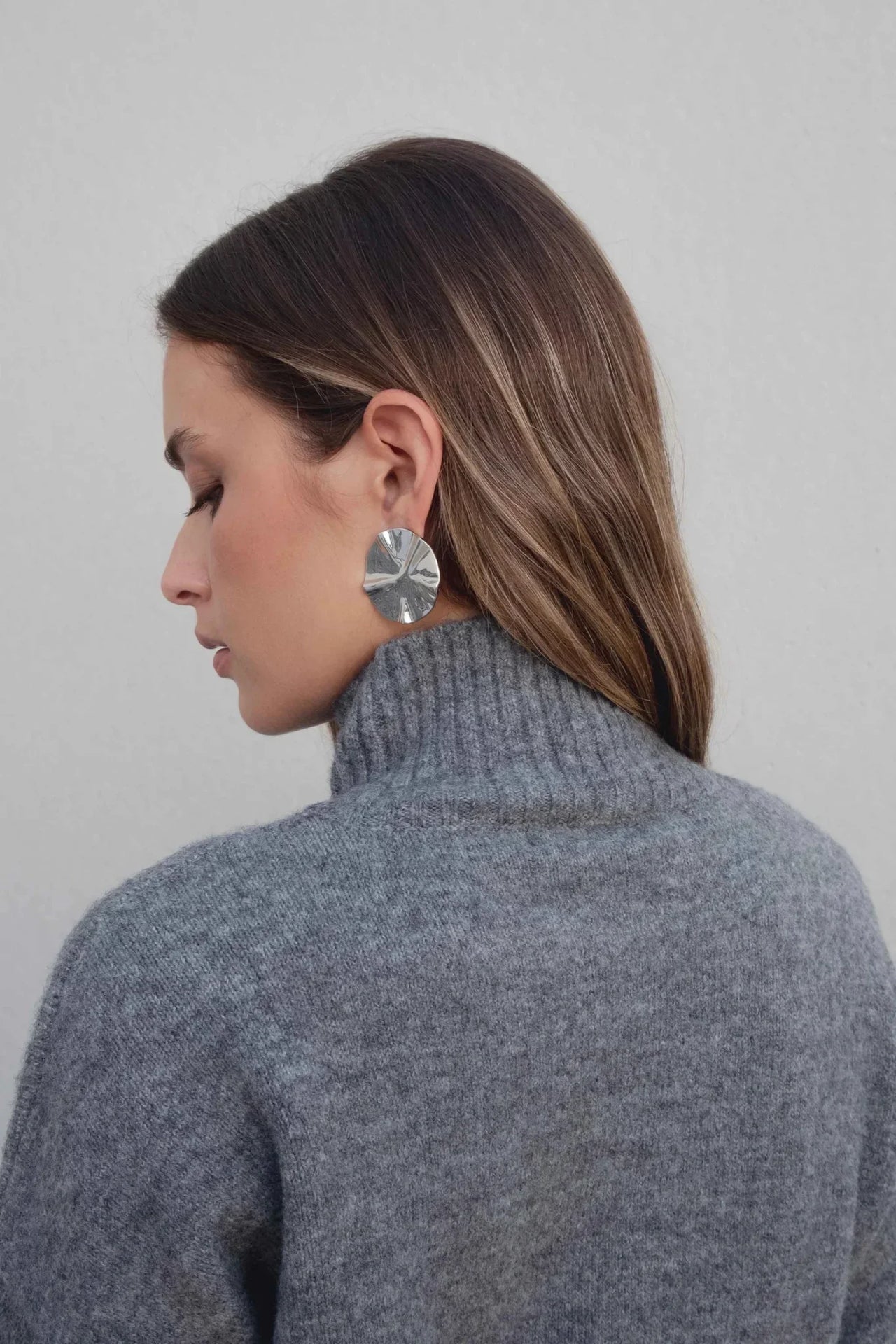 Femme de profil mettant en avant le pull col roulé femme gris et des boucles d'oreilles modernes.