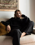 Pull Laine Femme Oversize à Manches Ballon - Femme détendue avec un pull laine femme noir, assise sur un canapé.