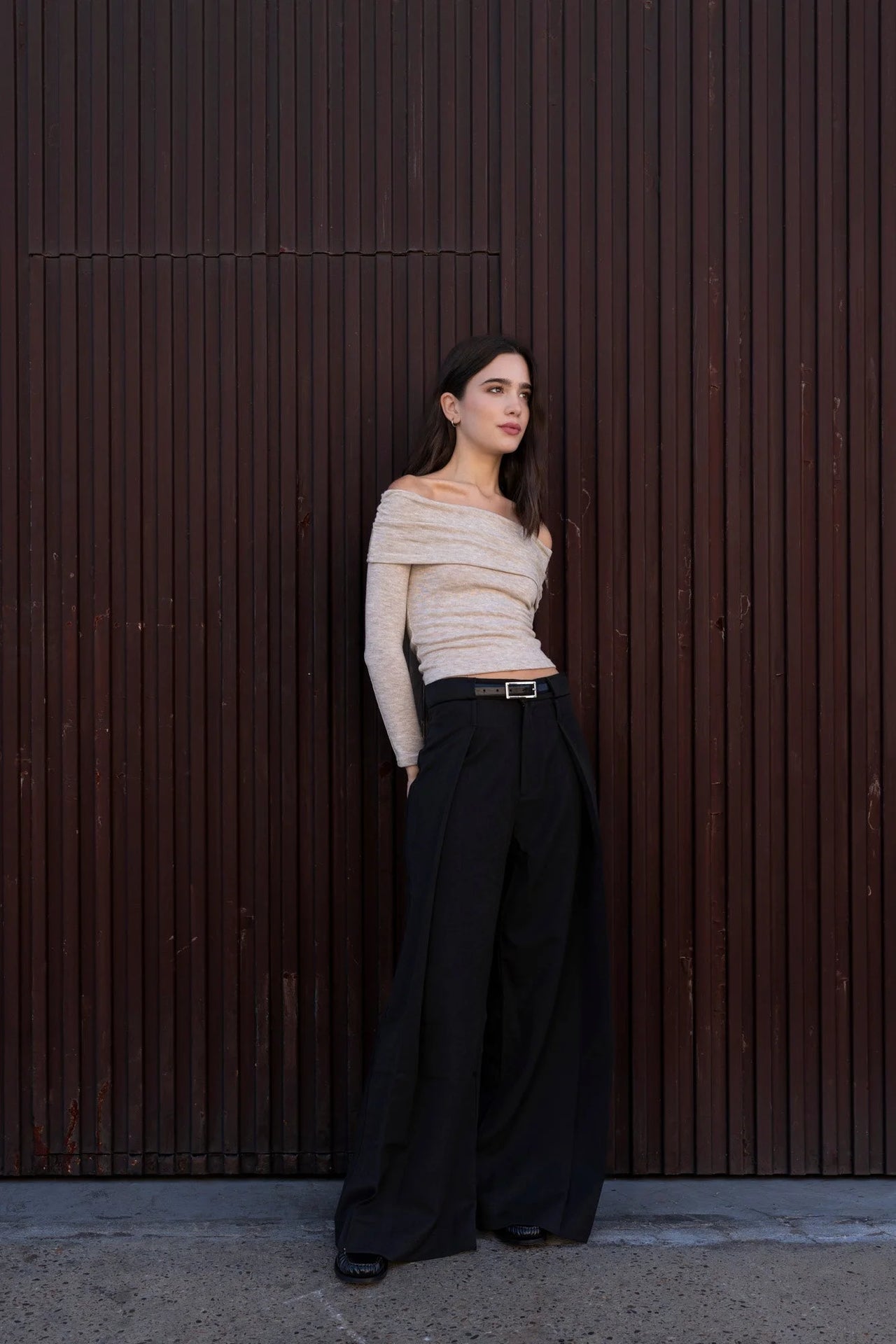 Jeune femme dans un pull épaule dénudée beige en tricot, se tenant devant un fond en bois, montrant la versatilité du style.