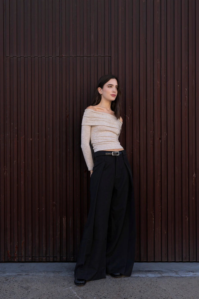 Jeune femme dans un pull épaule dénudée beige en tricot, se tenant devant un fond en bois, montrant la versatilité du style.