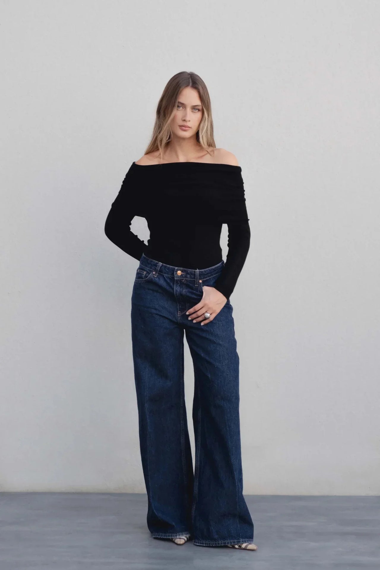 Pull femme maille noir à épaules dénudées, col bardot, porté par une femme debout avec un jean large.