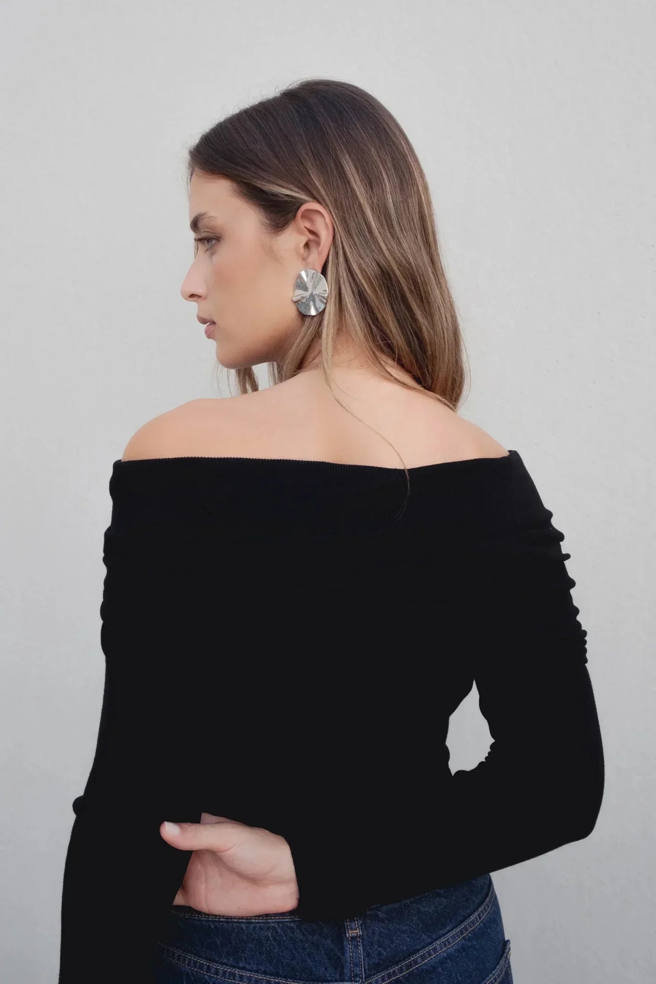 Femme vue de profil portant un pull femme maille noir à épaules dénudées, mettant en valeur son cou et ses boucles d'oreilles.