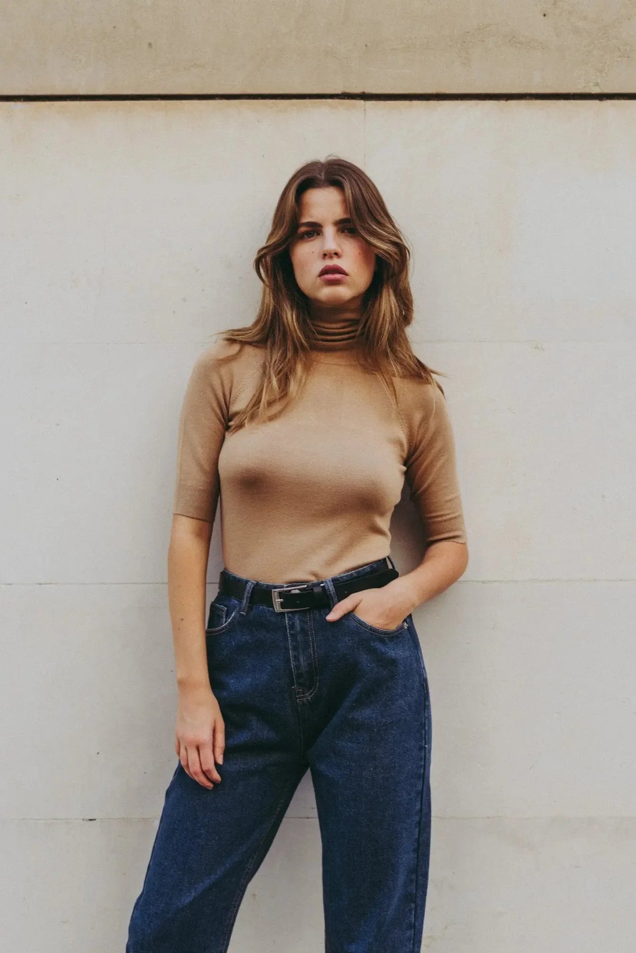 Pull col roulé femme beige tricoté, confortable et chic pour automne-hiver, modèle féminin tenant un pantalon en denim.