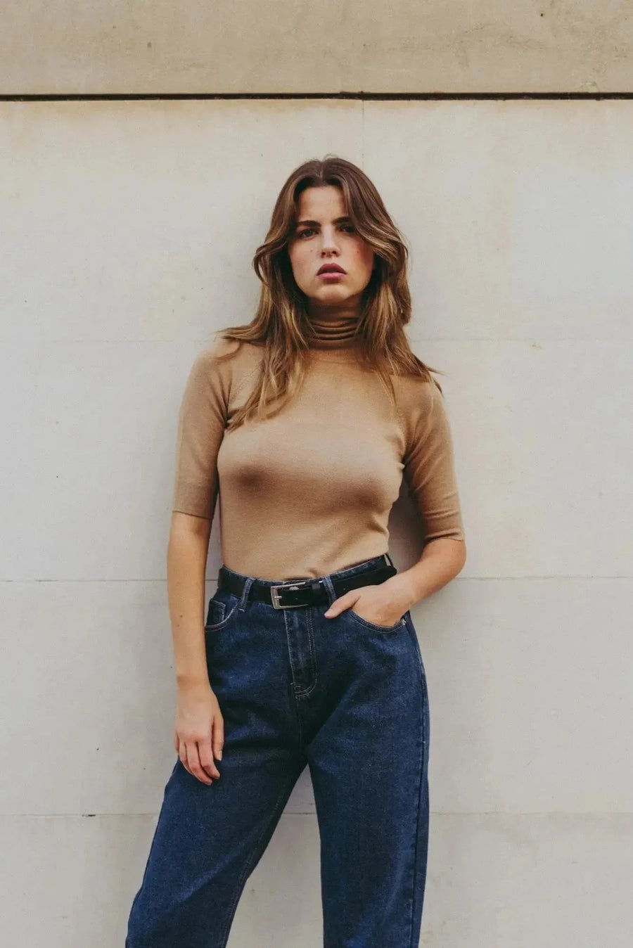 Pull col roulé femme beige tricoté, confortable et chic pour automne-hiver, modèle féminin tenant un pantalon en denim.