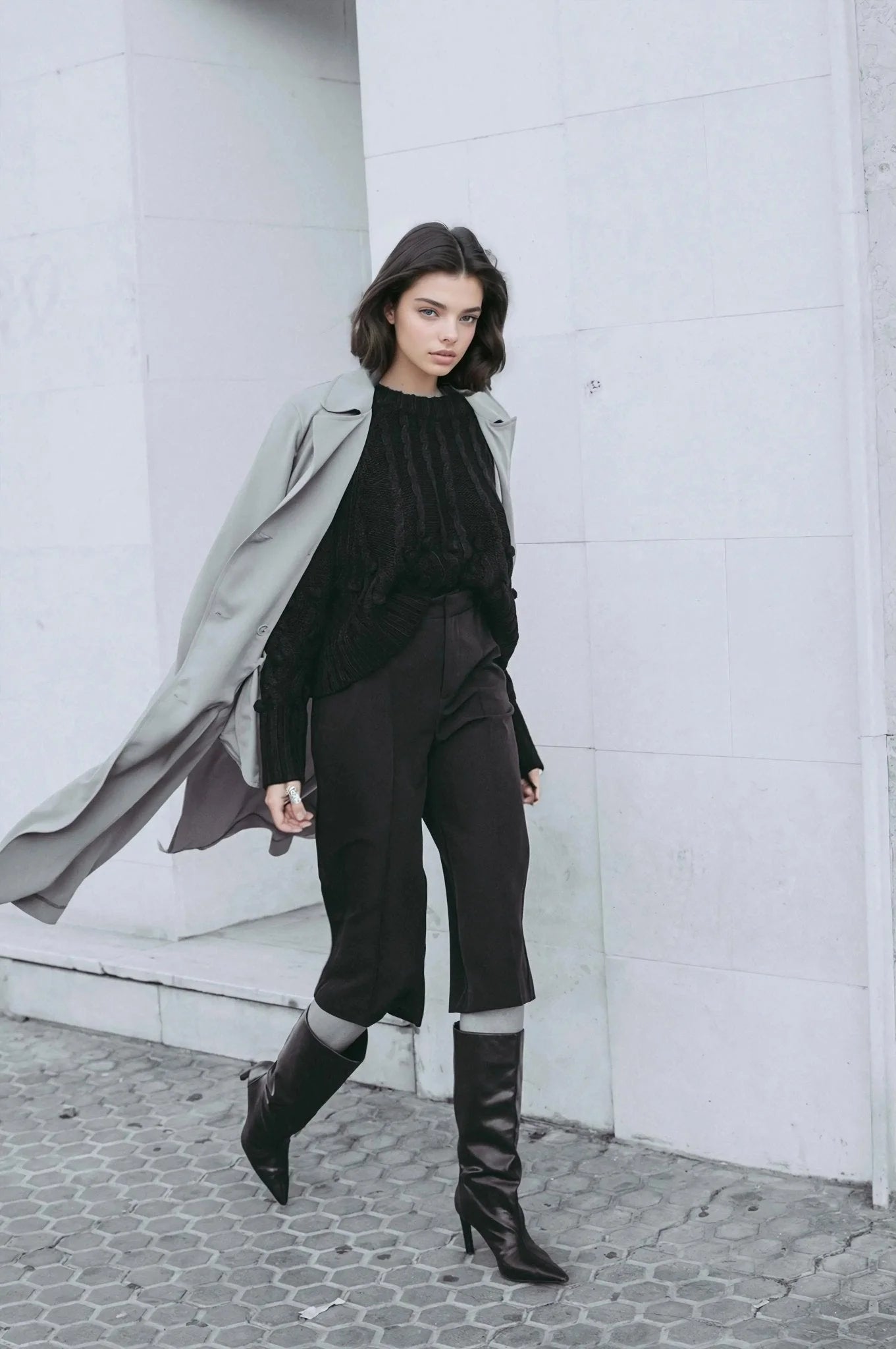 Femme en pull tricoté noir, pantalon court noir, bottes noires et trench gris clair