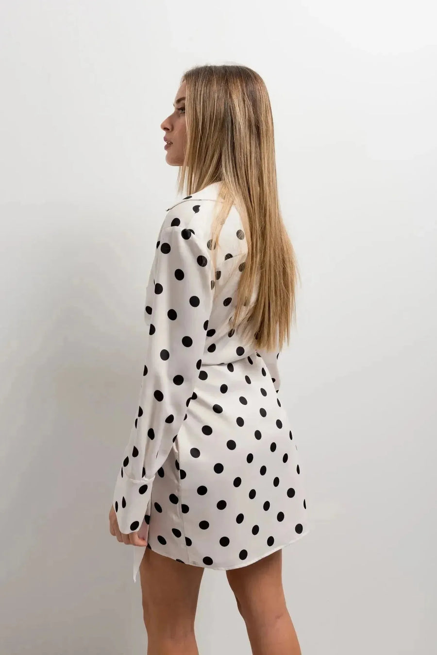 Femme de profil portant une robe à pois noir et blanc, mini robe portefeuille élégante à manches longues, montrant le dos de la robe.