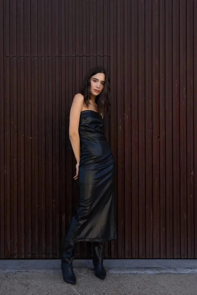 Femme stylée en robe longue simili cuir noir bustier midi avec fente latérale - Élégance Audacieuse, avec des bottes pour un look tendance.