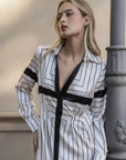 Femme portant une robe chemise longue chic à rayures noires et blanches, posant élégamment contre un pilier, parfaite pour un look élégant et confortable.