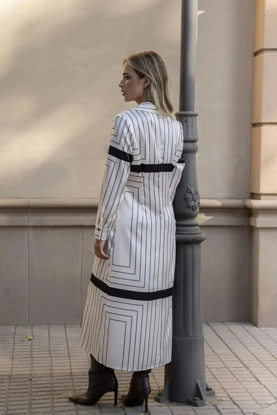 Femme vue de dos dans une robe chemise longue à rayures noires et blanches, s'appuyant contre un pilier, pour un look sophistiqué.