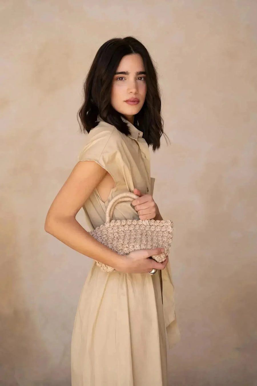 Profil d'une femme avec une robe chemise femme beige midi ceinturée, tenant un petit sac en paille, illustrant le style chic et décontracté.
