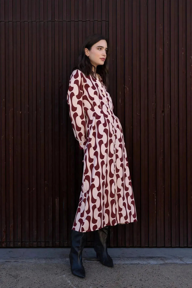 Femme stylée en robe chemise longue imprimée bordeaux, avec un look décontracté, posant légèrement de côté.