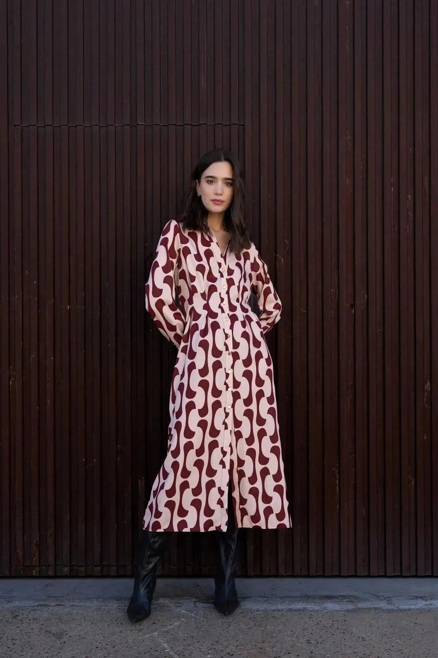 Femme portant une robe chemise longue imprimée bordeaux avec un style fluide, boutons dorés et col chemise, posant devant un fond de mur en bois.