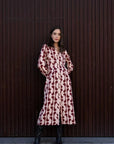Femme portant une robe chemise longue imprimée bordeaux avec un style fluide, boutons dorés et col chemise, posant devant un fond de mur en bois.