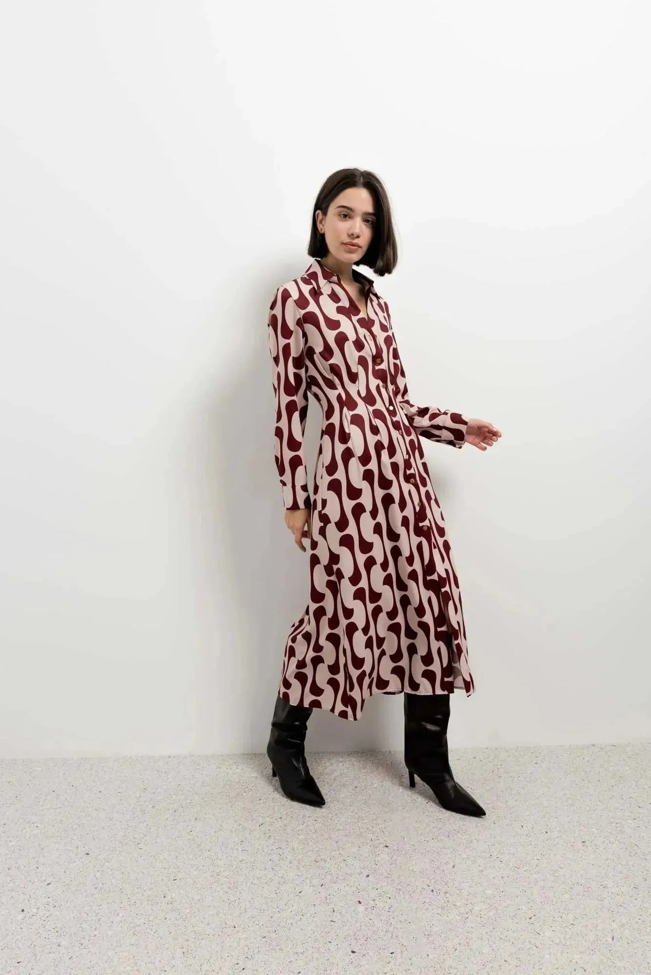 Femme en robe chemise longue imprimée bordeaux prenant une pose dynamique, avec des bottes noires dans un intérieur moderne.