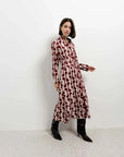 Femme en robe chemise longue imprimée bordeaux prenant une pose dynamique, avec des bottes noires dans un intérieur moderne.