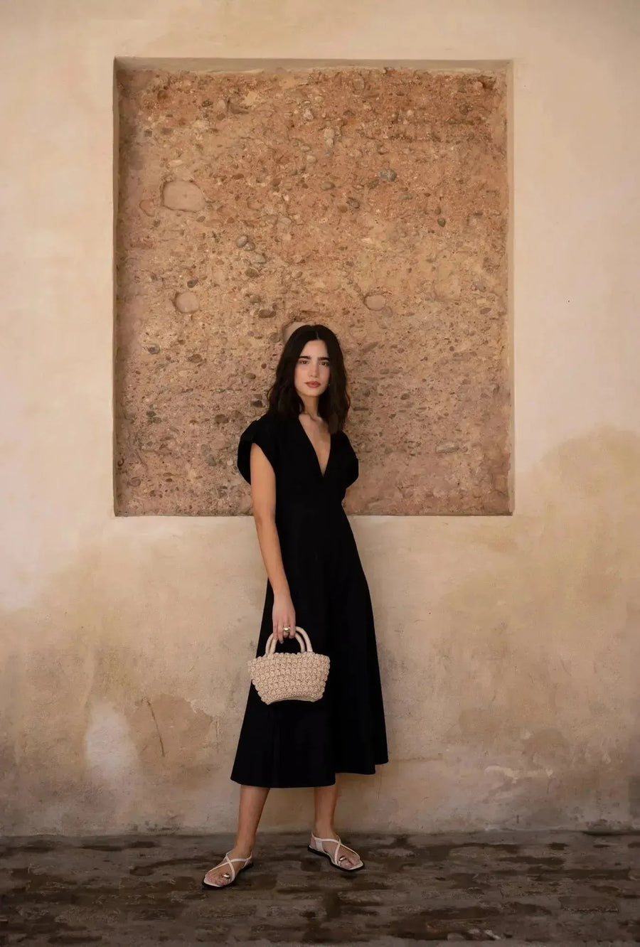 Femme élégante avec une robe popeline noire chic à manches courtes - décolleté en V évasée pour toutes occasions, à côté d'un mur beige.