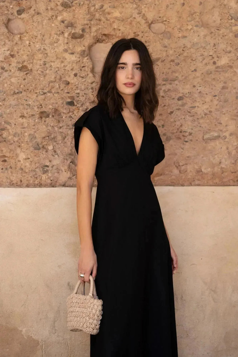 Femme portant une robe popeline noire chic à manches courtes - décolleté en V évasée pour toutes occasions, posant devant un mur en pierre.