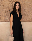 Femme portant une robe popeline noire chic à manches courtes - décolleté en V évasée pour toutes occasions, posant devant un mur en pierre.