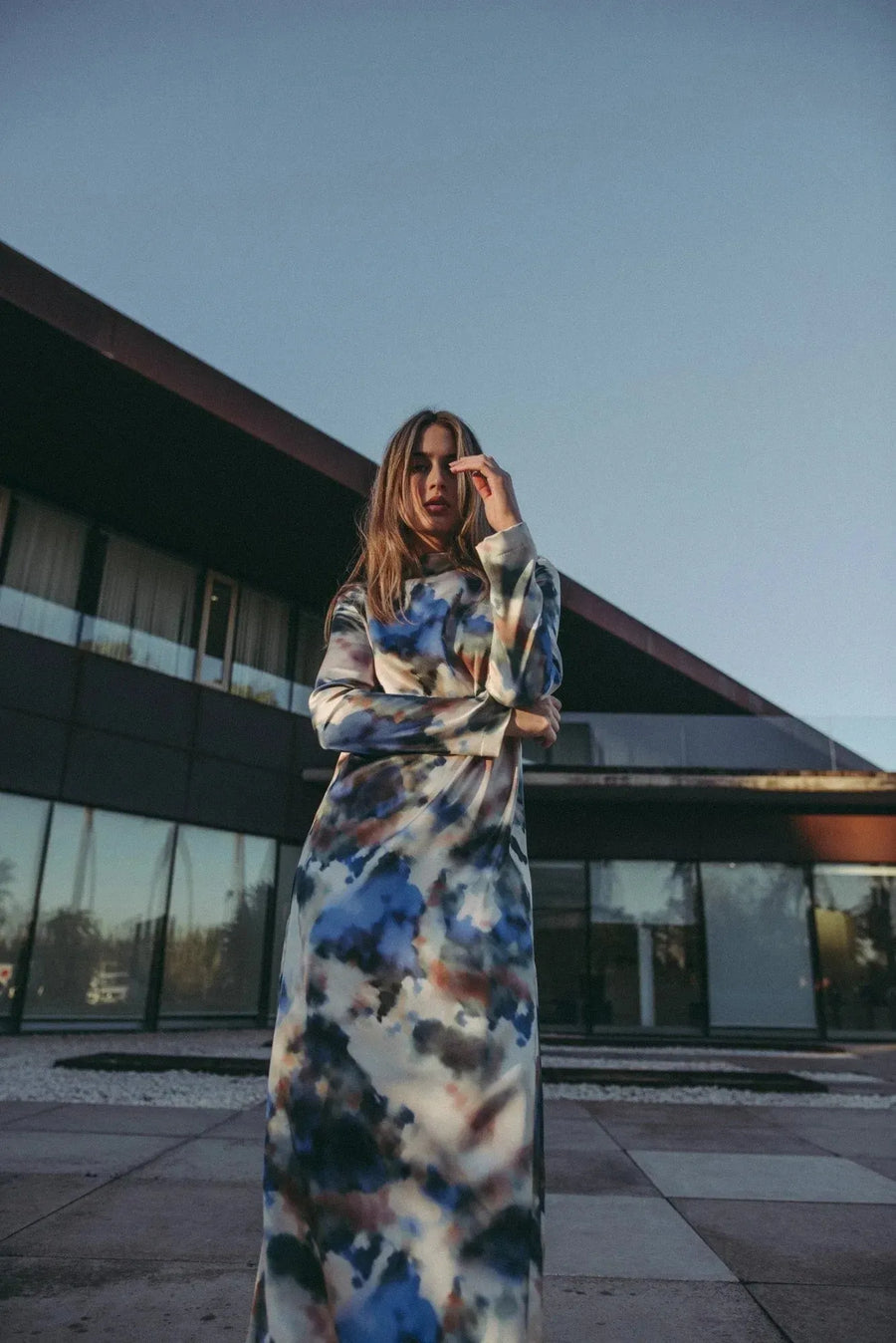 Femme posant avec assurance dans une robe satin longue manche longue aux motifs aquarelle, devant un design architectural contemporain, incarnant le style et le confort.
