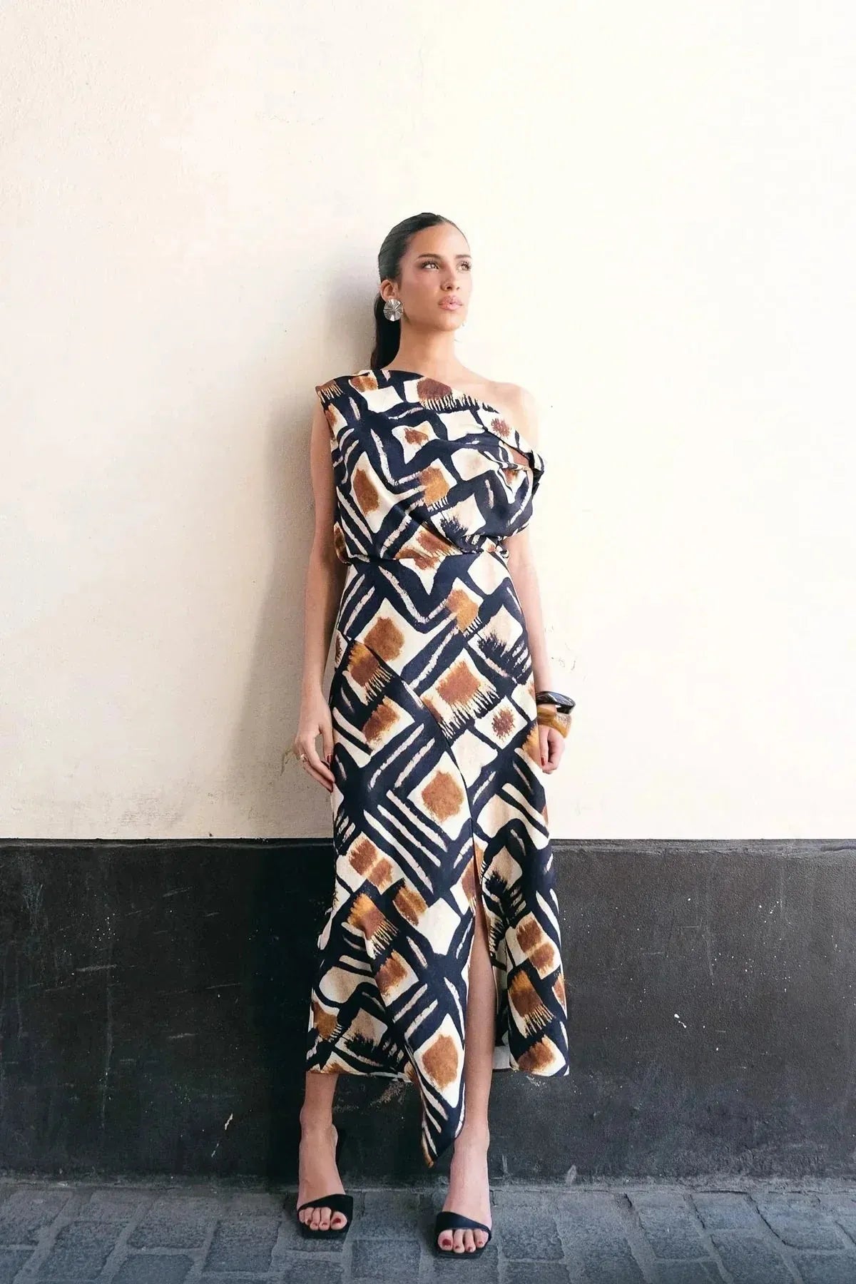 Une femme en haut femme asymétrique à une épaule imprimé graphique marron, noir et blanc, posant contre un mur. Ce style chic et moderne est parfait pour diverses occasions.