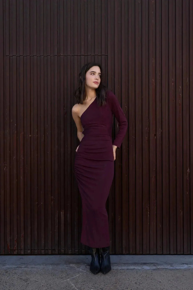 Femme affichant la robe asymétrique une manche bordeaux devant un mur texturé, mettant en avant son style unique.