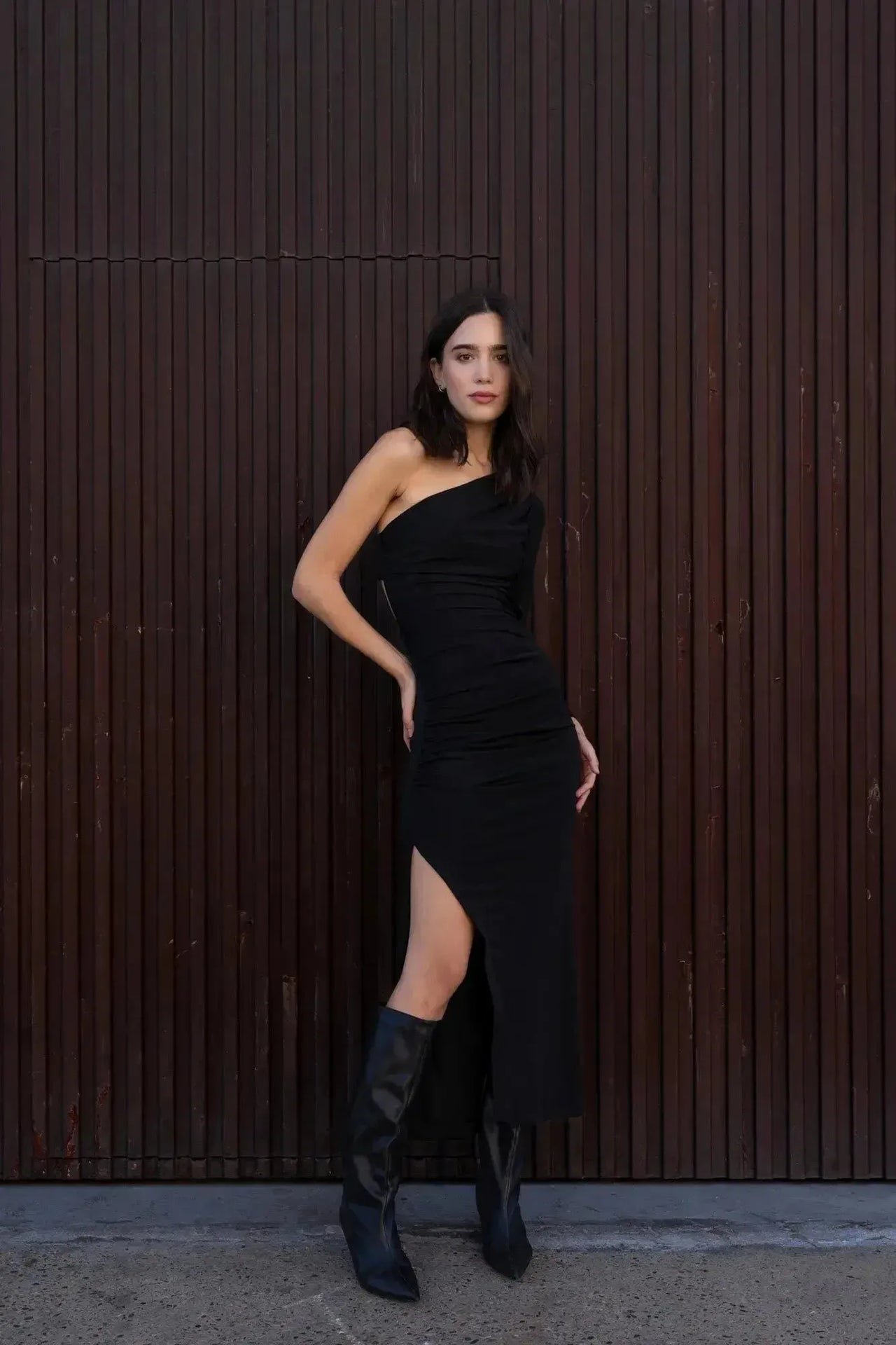 Modèle portant une robe asymétrique une manche noir chic, élégante et confortable pour soirées et mariages, avec des bottes noires en cuir.