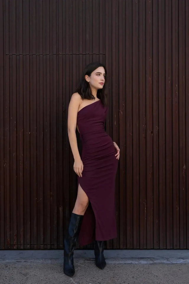 Modèle en robe asymétrique une manche bordeaux, captivant le regard avec une pose gracieuse près d'un mur.