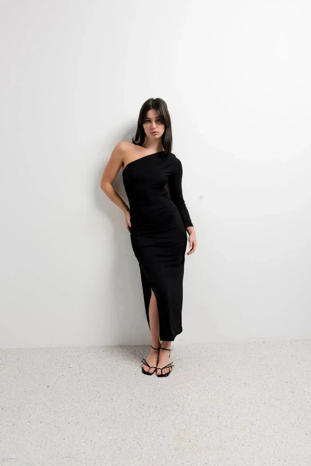 Femme en robe asymétrique une manche noire, au style élégant, posant devant un mur blanc pour mettre en avant la élégance de la robe.