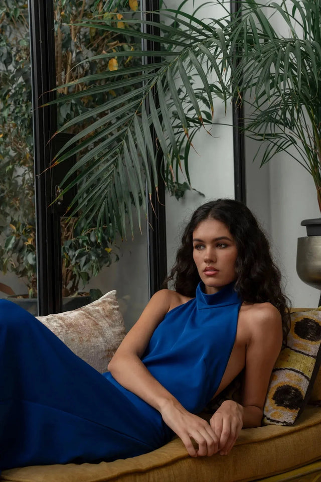 Model assise dans une robe longue dos nu Sapiato bleue, dans un environnement intérieur, marquant son allure chic et décontractée.