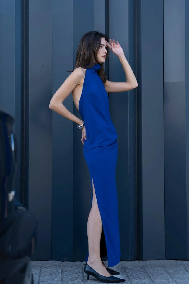 Modèle en robe longue dos nu Sapiato de couleur bleue, affichant un style chic, se tenant de profil devant une porte moderne.