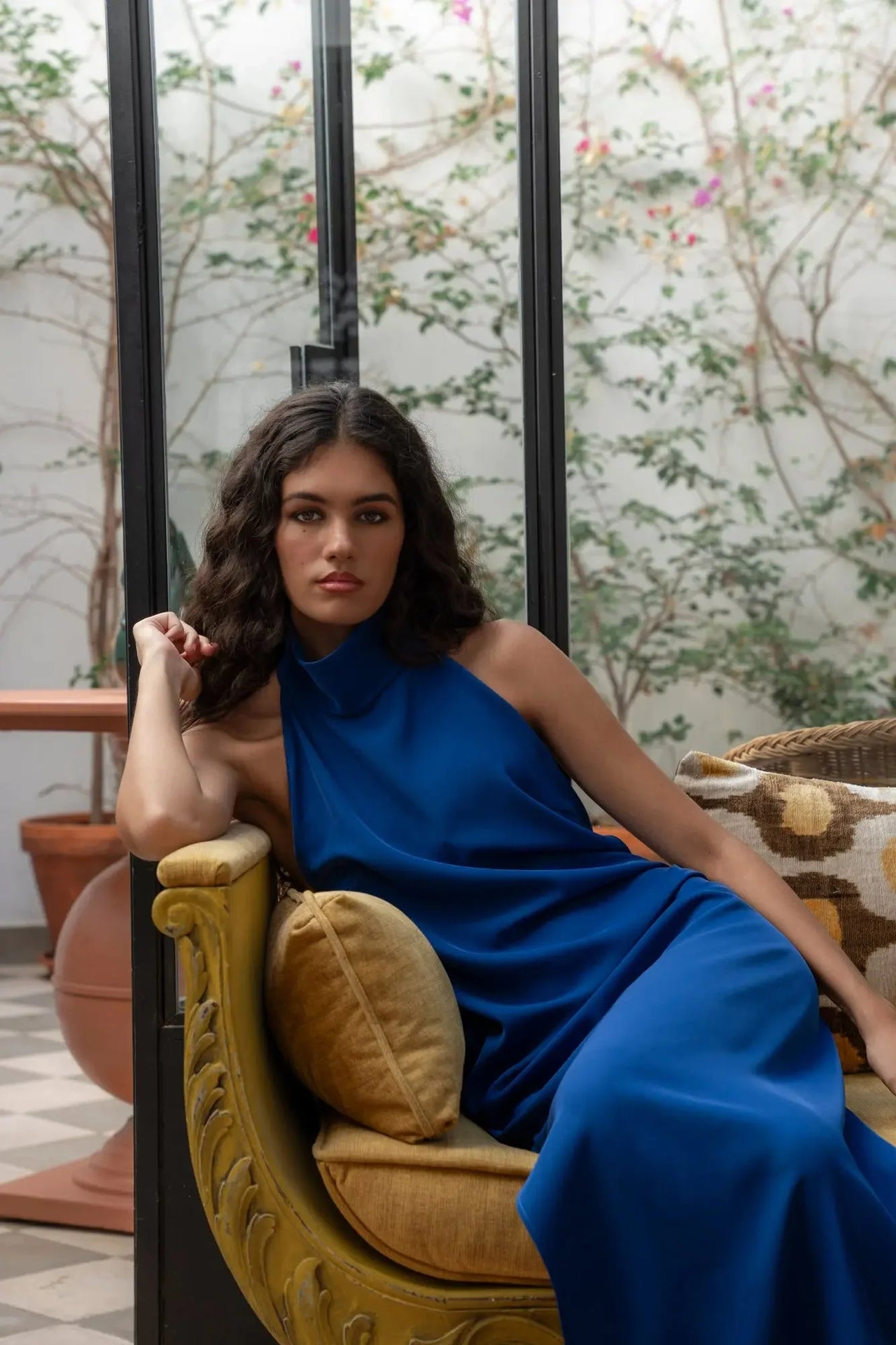 Femme dans une robe longue dos nu Sapiato, posant sur un canapé, l'air chic et détendu dans un cadre artistique.