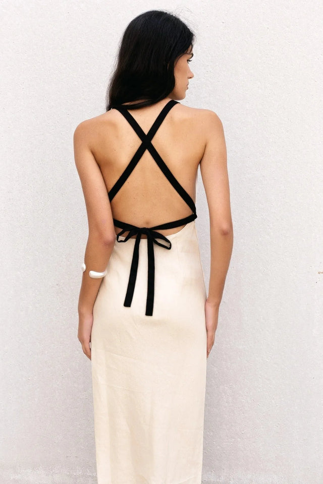 Détails du dos d'un modèle portant une robe longue dos nu en lin écru, montrant les bretelles croisées noires et le nouage, idéale pour un look chic.