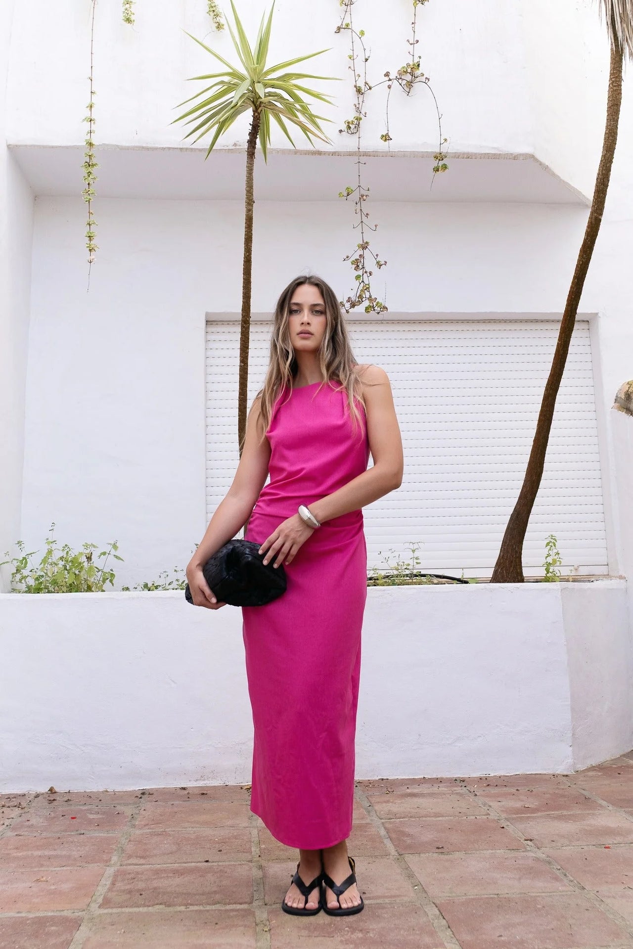 Modèle debout dans une robe fuchsia longue chic en lin, sans manches, encolure froncée, style minimaliste, posé dans un cadre extérieur.
