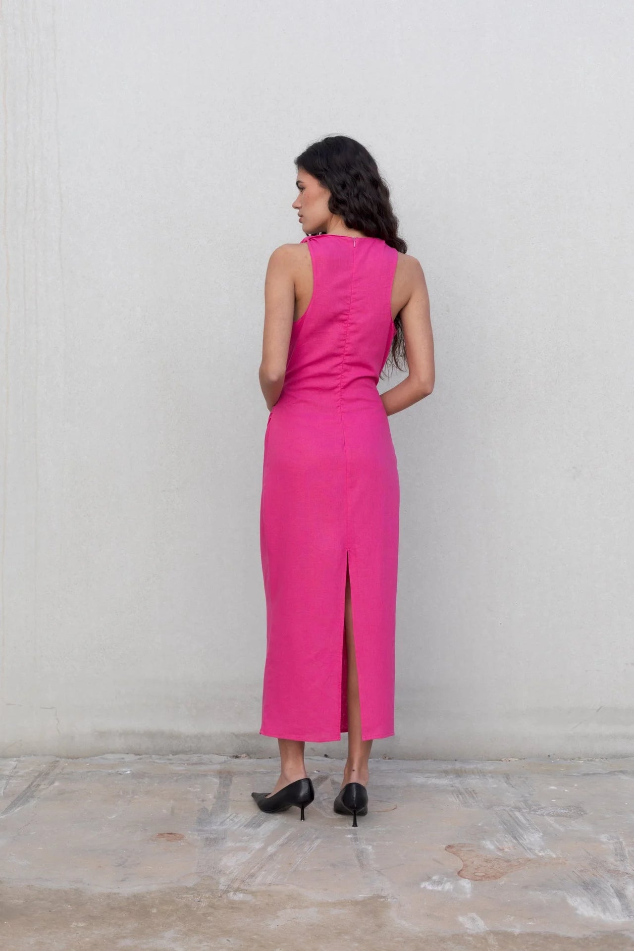 Femme vue de dos dans une robe fuchsia longue chic en lin, sans manches, encolure froncée, style minimaliste, révélant une fente.