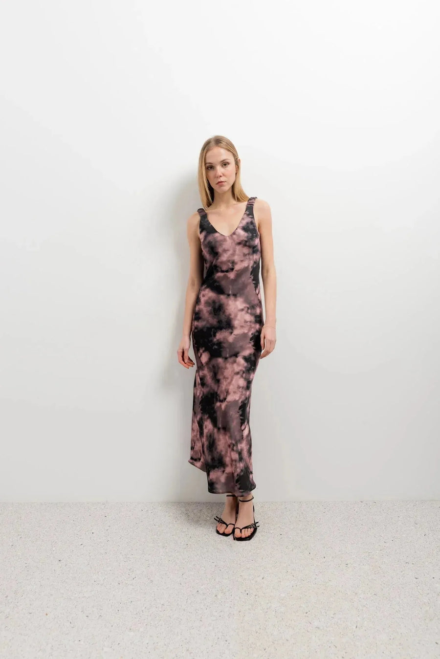 Femme en robe tie dye longue satinée noir et rose, assise avec des sandales pour une touche décontractée.