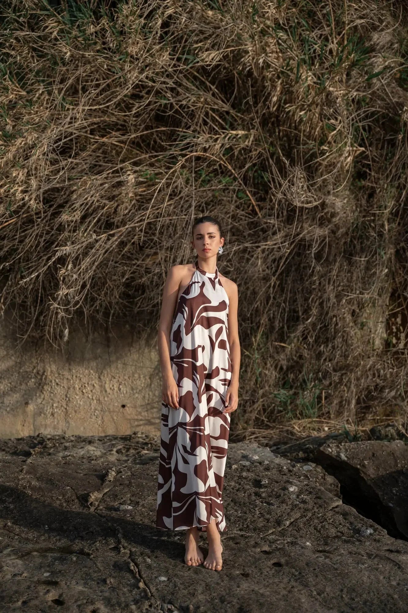 Robe Longue Bohême Imprimée Chocolat & Blanc Dos Nu Fluide avec Col Licou, femme posant debout près d'un rocher, entourée de végétation sauvage.