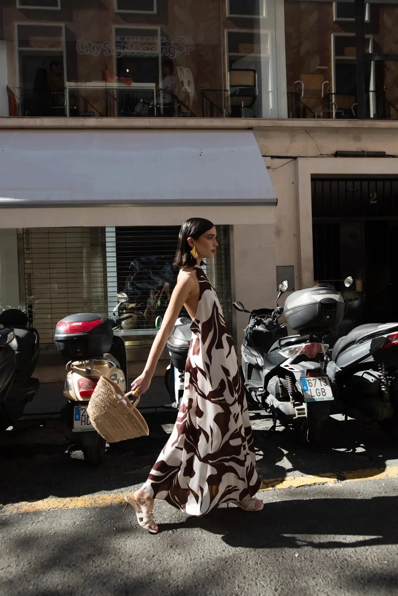 Robe Longue Bohême Imprimée Chocolat & Blanc Dos Nu Fluide avec Col Licou, présentée par une femme marchant dans la rue, style décontracté avec un sac en osier.