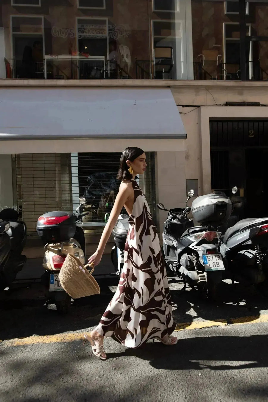 Robe Longue Bohême Imprimée Chocolat & Blanc Dos Nu Fluide avec Col Licou, présentée par une femme marchant dans la rue, style décontracté avec un sac en osier.
