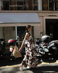 Robe Longue Bohême Imprimée Chocolat & Blanc Dos Nu Fluide avec Col Licou, présentée par une femme marchant dans la rue, style décontracté avec un sac en osier.