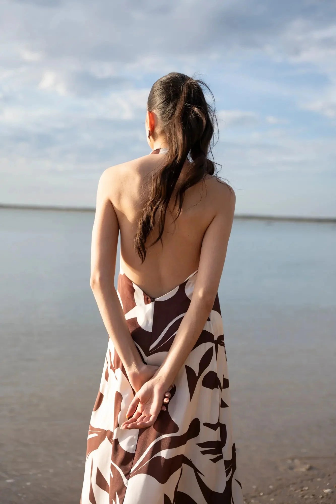 Robe Longue Bohême Imprimée Chocolat & Blanc Dos Nu Fluide avec Col Licou, vue arrière du modèle montrant le dos nu, au bord de la mer.