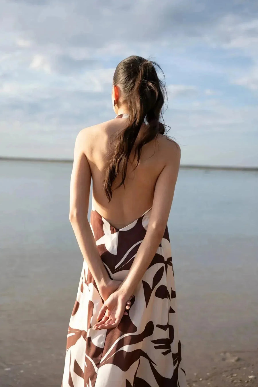 Robe Longue Bohême Imprimée Chocolat & Blanc Dos Nu Fluide avec Col Licou, vue arrière du modèle montrant le dos nu, au bord de la mer.